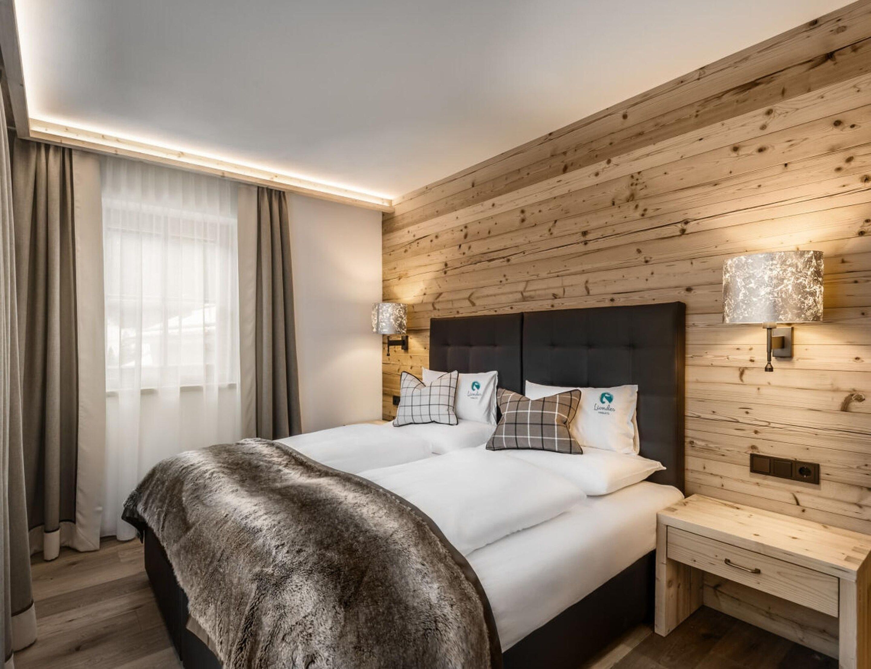 A modern hotel room with a wooden wall panel, a large bed with white bedding, two bedside lamps, and a small bedside table.