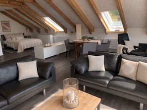 A spacious loft with a high ceiling, exposed wooden beams, a central seating area with leather sofas, a dining table, and a view through a skylight. - Holiday Apartment With 1 Bedroom (Gluecksburg)