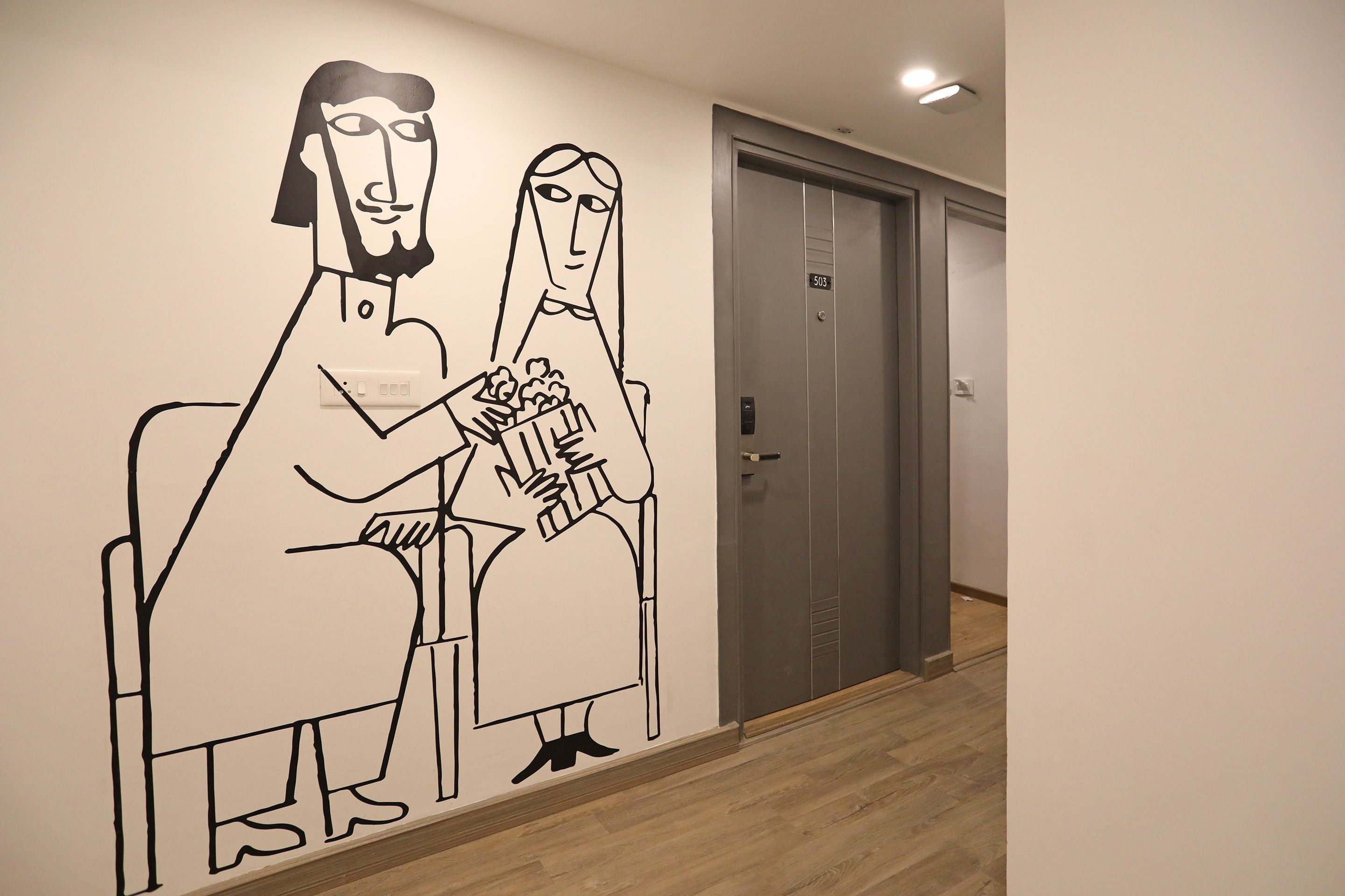 Un mur orné d’une fresque représentant deux personnes assises, une porte avec une plaque numérotée et un luminaire fixé au plafond.