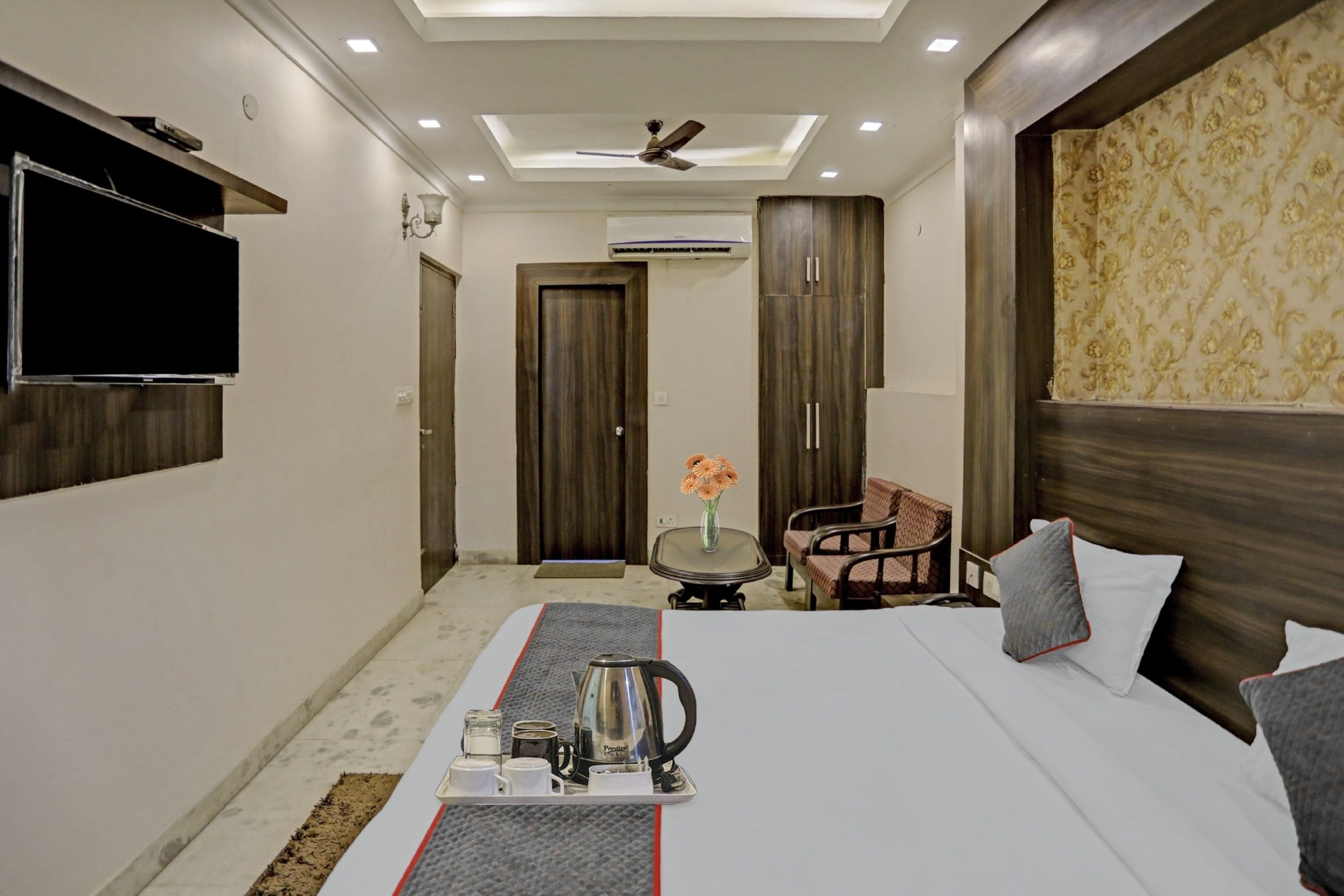 A hotel room with a bed, a TV mounted on the wall, a ceiling fan, and a small table with a vase of flowers.