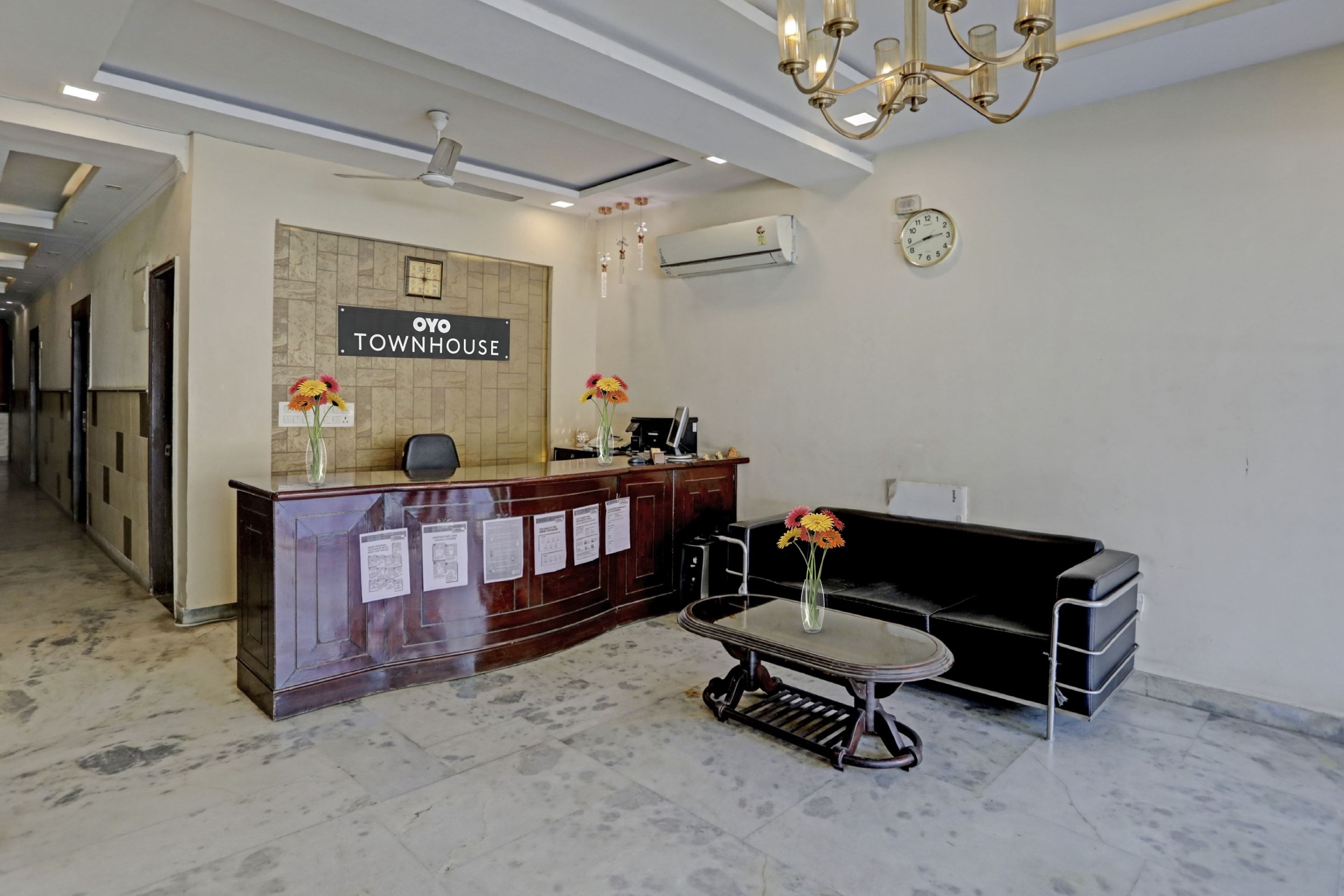 Un espace d’accueil avec une enseigne indiquant « OYO TOWNHOUSE », un comptoir de réception, un canapé noir, une petite table sur laquelle est posé un vase de fleurs et une horloge au mur.