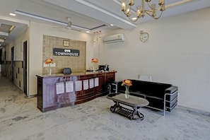 Zona de recepción con un letrero que dice 'OYO TOWNHOUSE', un mostrador de recepción, un sofá negro, una pequeña mesa con un jarrón de flores y un reloj en la pared.