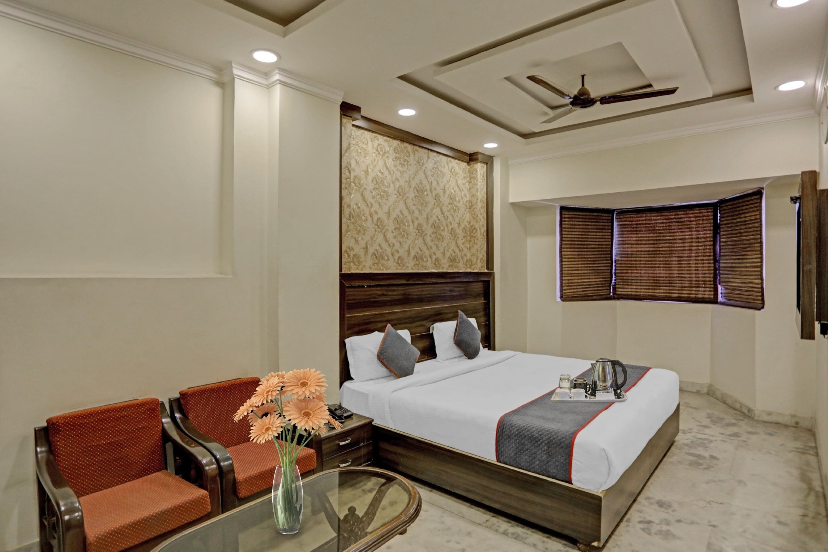 Habitación de hotel con cama, zona de estar con sillas, una pequeña mesa con un jarrón de flores y un ventilador de techo.