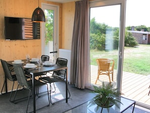 Ein Essbereich mit Tisch für vier Personen, einem an der Wand befestigten Fernseher und Blick auf einen Garten durch eine Schiebetür.