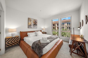 A hotel room with a bed, bedside tables, a desk, and a window with a view of buildings. - Chic Resort-Style 2BR - Pools, BBQ & Balcony (Lake Forest)