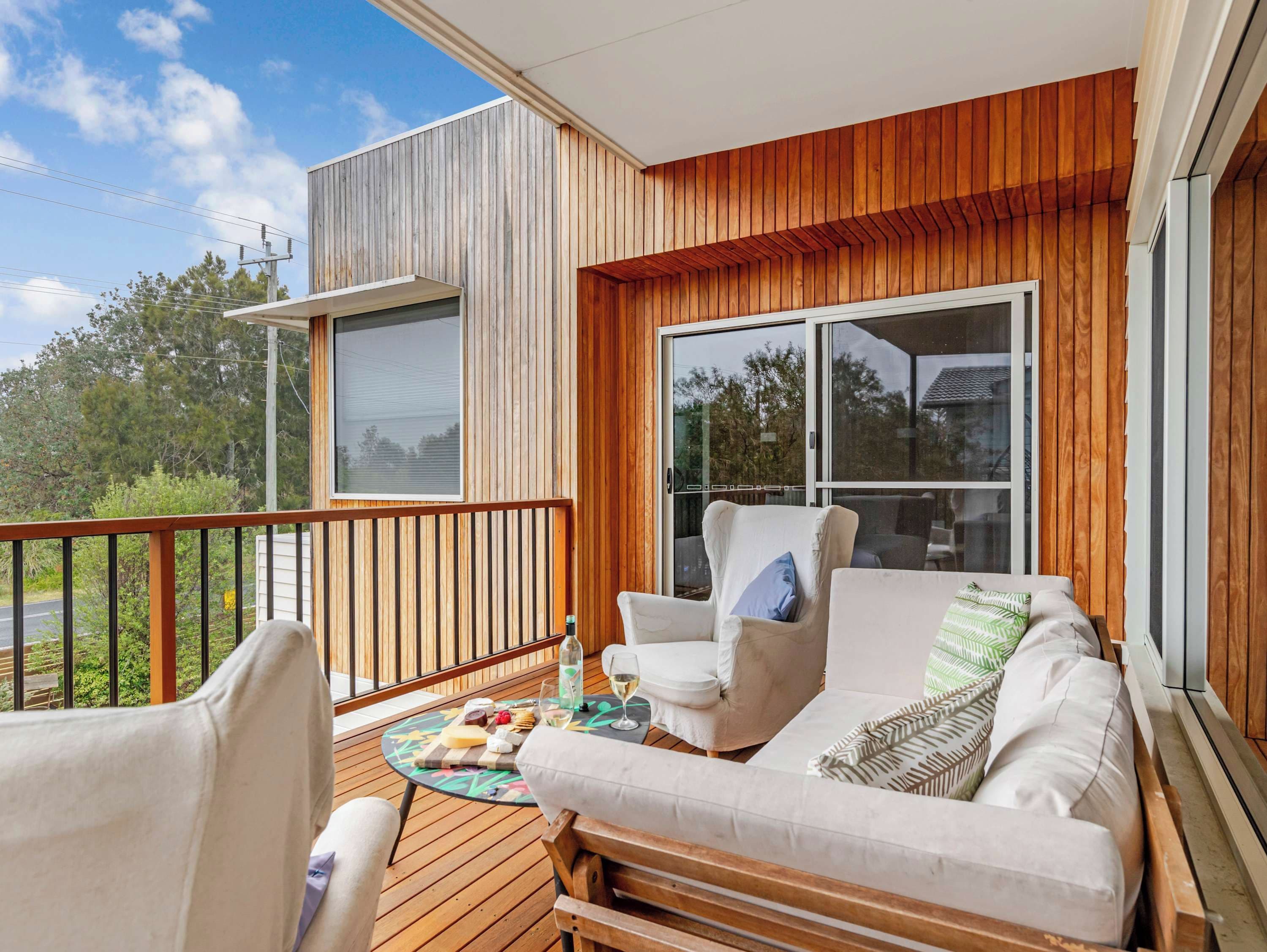 A balcony with a wooden deck, a sofa, two armchairs, and a small table with a cheese board and drinks.