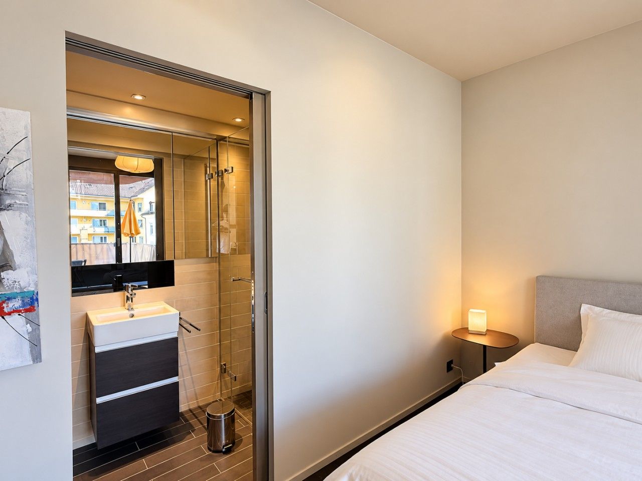 Quarto de hotel com cama, mesa de cabeceira com luminária, banheiro com pia e chuveiro, e vista para o exterior.