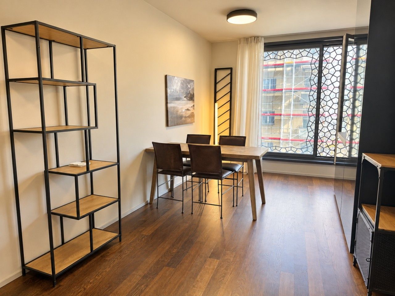 Une pièce comprenant une table à manger, des chaises, une bibliothèque et une fenêtre donnant sur des immeubles.