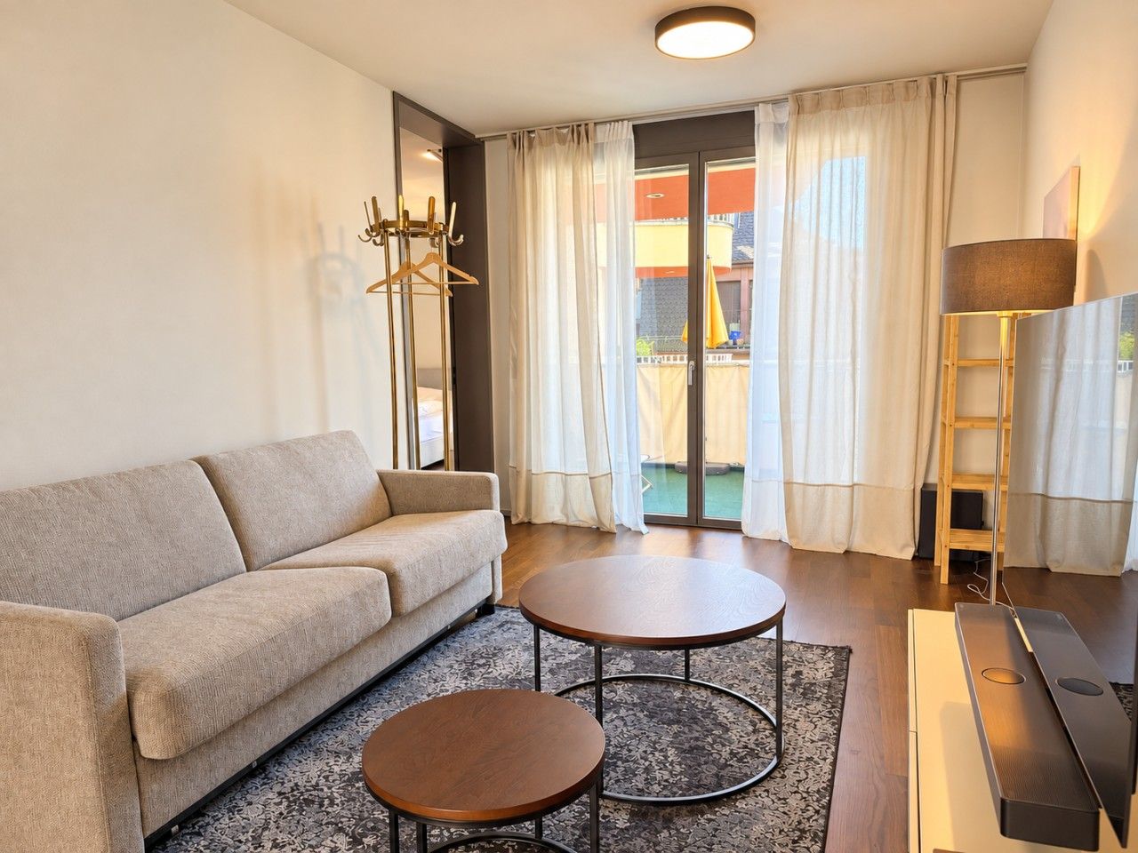 Ein modernes Wohnzimmer mit einer Couch, zwei runden Tischen, einem Fernseher und einem Balkon mit Vorhängen.