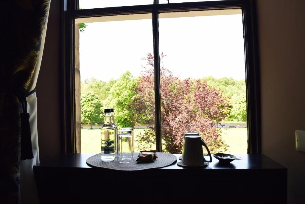 Una mesa con una botella, un vaso y una jarrón junto a una ventana con vistas a árboles.