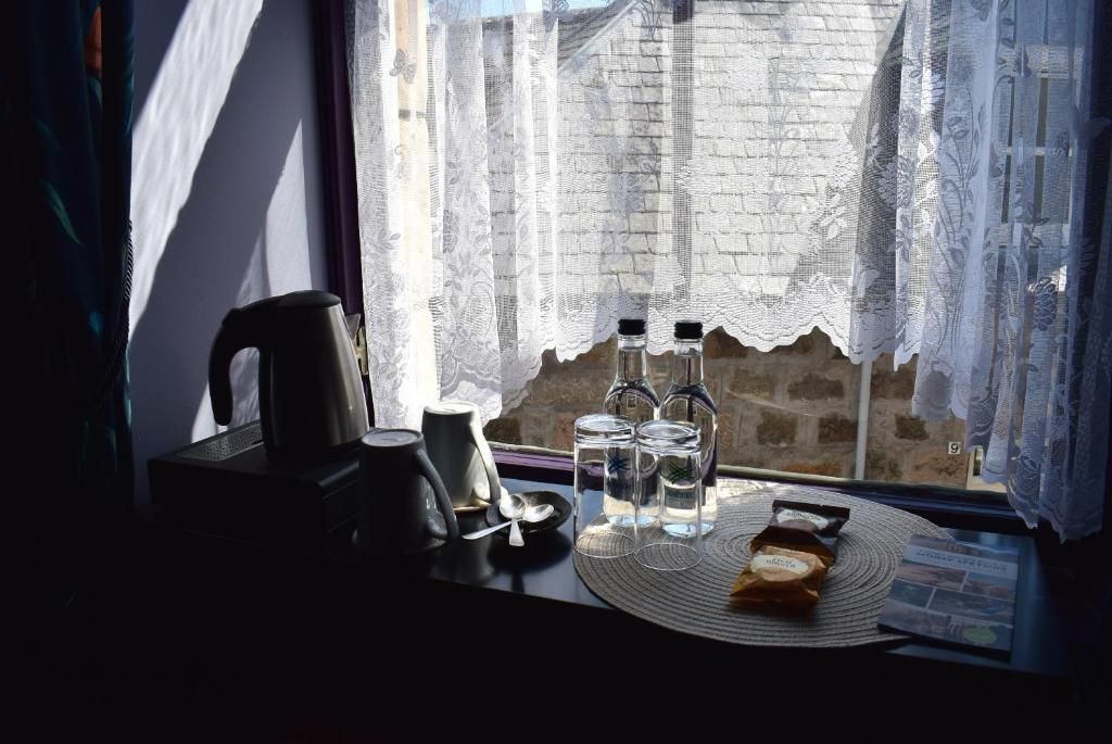 A table with a kettle, cups, and bottles, near a window with lace curtains.