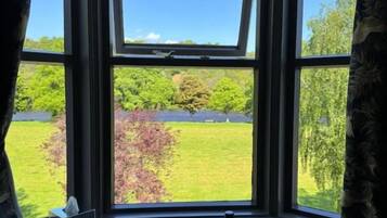 Una habitación con ventanal que da a un paisaje verde, una mesa con una botella, vasos y un plato, y una ventana con cortina.