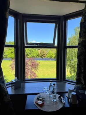 A room with a bay window overlooking a green landscape, a table with a bottle, glasses, and a plate, and a window with a curtain. - The Mash Tun (Charlestown of Aberlour)