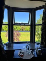 Una habitación con ventanal que da a un paisaje verde, una mesa con una botella, vasos y un plato, y una ventana con cortina.