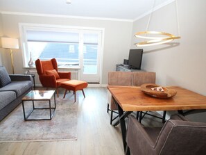 A modern living room with a sofa, armchair, ottoman, coffee table, dining table, and a television. - The Beach House Holiday Apartment 2 (Sylt)