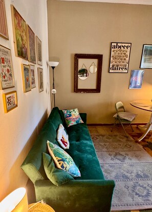 Ein Wohnzimmer mit einem grünen Sofa, einer Stehlampe, einem gemusterten Teppich und verschiedenen gerahmten Kunstwerken an den Wänden.