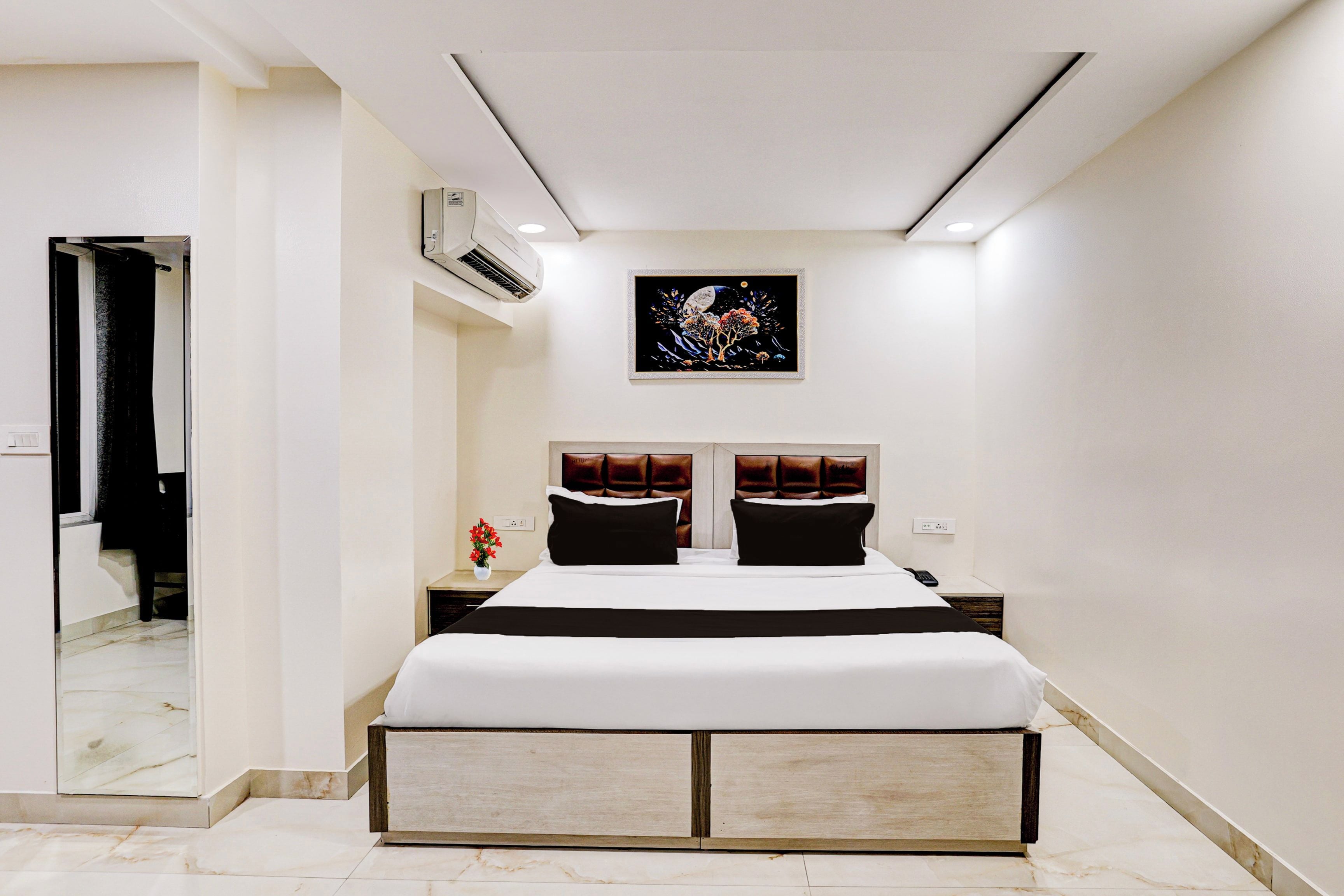 Une chambre moderne avec un grand lit, une table de chevet, un climatiseur fixé au mur et une œuvre d’art encadrée au-dessus du lit.