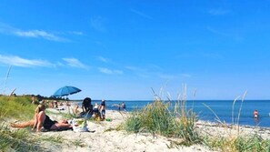 Une plage où des gens se prélassent au soleil et se baignent, un parasol bleu et de l’herbe au premier plan.
