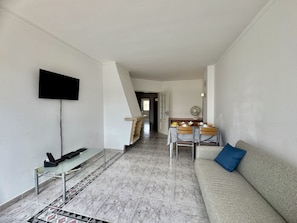 Ein Wohnzimmer mit Flachbildfernseher, einer Couch, einem Esstisch mit Stühlen und einer Treppe zu einem weiteren Raum.