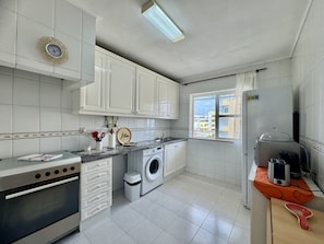 A compact kitchen with white cabinets, a washing machine, a refrigerator, and a window with a view of buildings. - Faro Central 4 by Homing (Faro)