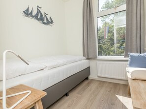 Ein kleines, ordentlich eingerichtetes Schlafzimmer mit einem Einzelbett, einem Nachttisch, einem Fenster mit Vorhängen und einem Wandbild mit Segelbooten.