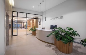 A modern reception area with a reception desk, a large potted plant, and a glass door entrance leading to a waiting area. - Mburucuya 1BR | Rooftop Pool, Gym & Parking (Asunción)