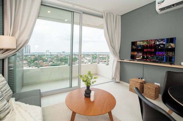 Ein modernes Wohnzimmer mit einer großen Schiebeglastür, einem runden Couchtisch aus Holz, einem Blumentopf, einem flachbildfernseher an der Wand und Blick auf eine Stadtlandschaft.