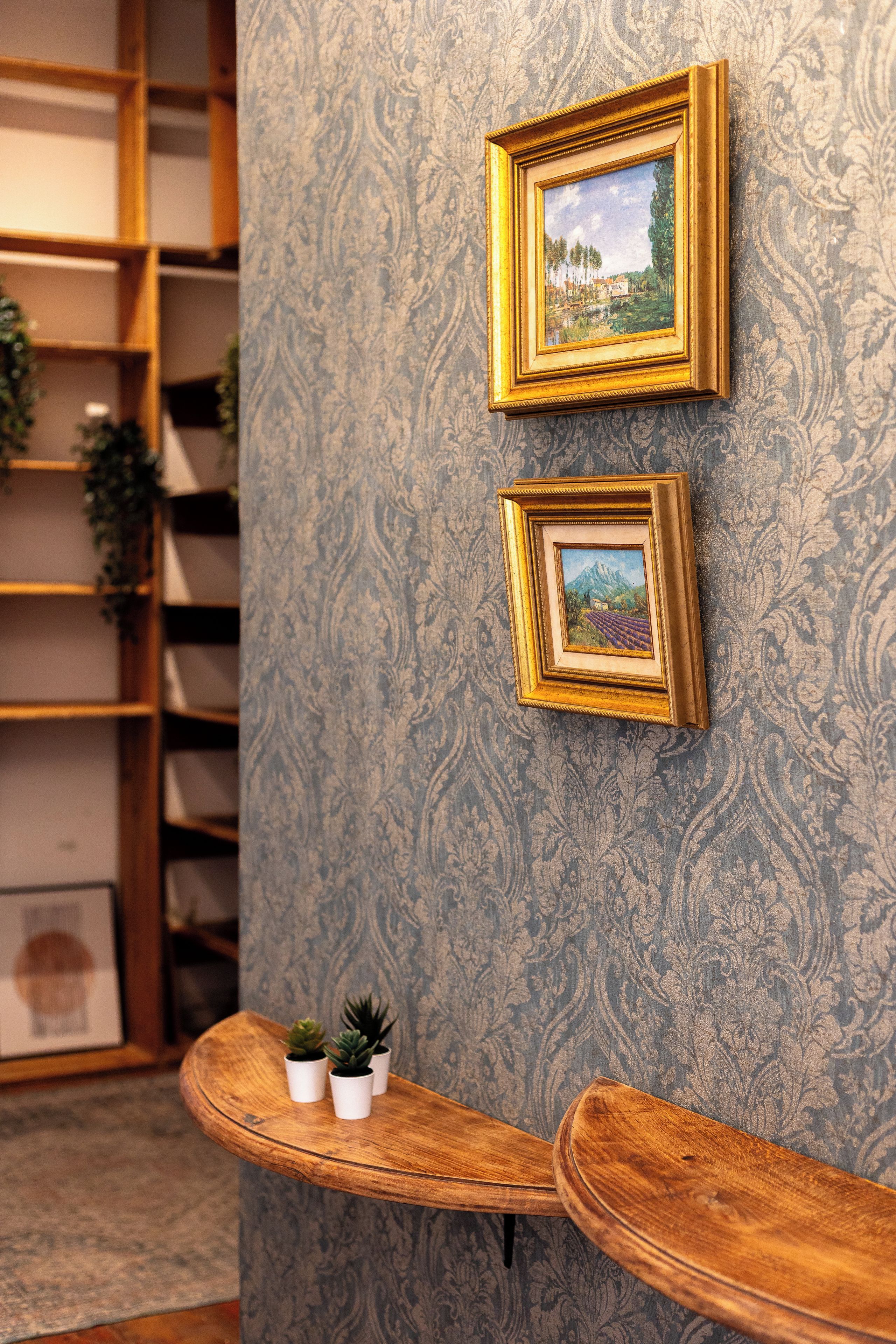 Una habitación con dos cuadros en la pared, un banco de madera con plantas en macetas y un papel tapiz estampado.