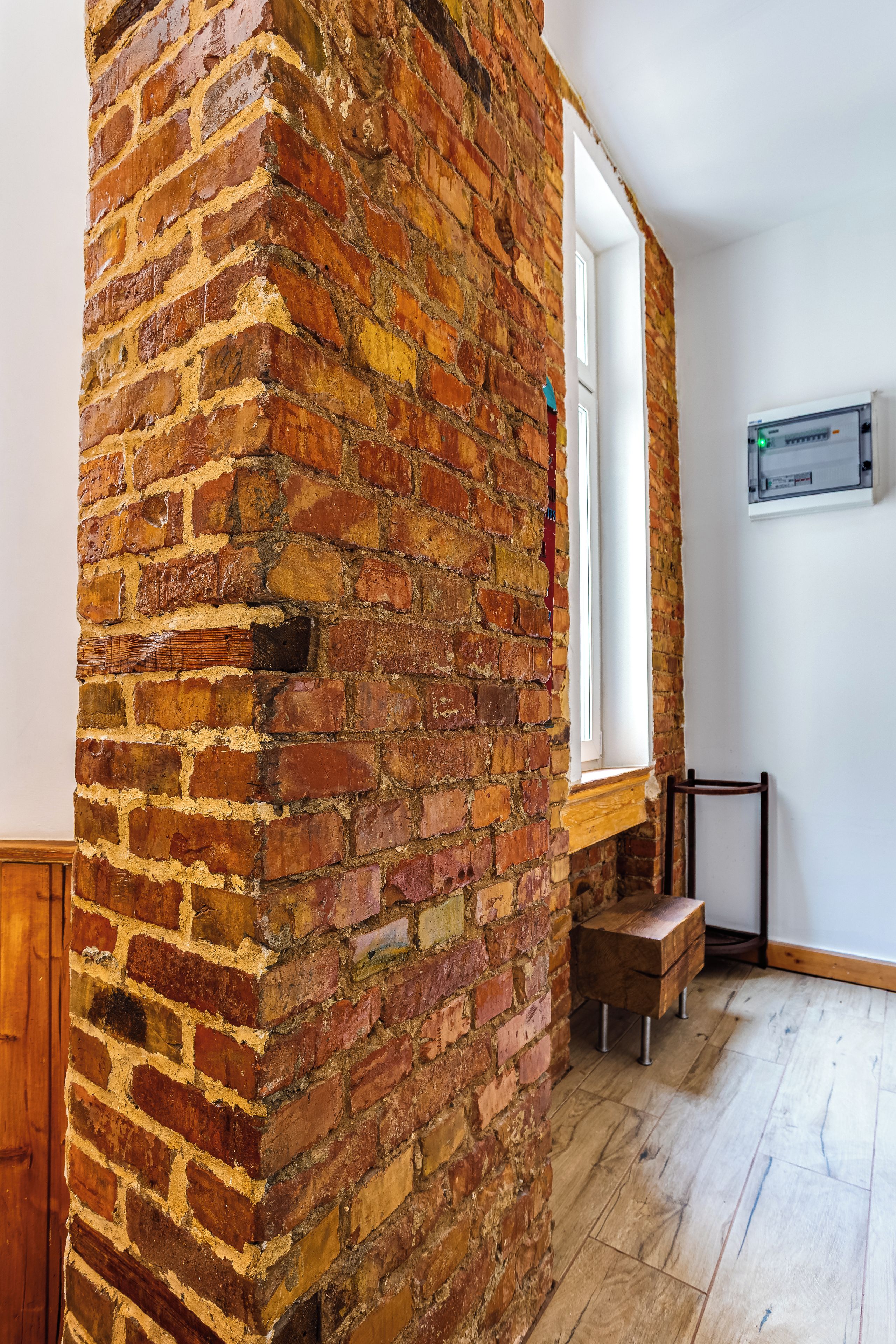 A room with a brick wall, a window, a wooden bench, and a power outlet on the wall.