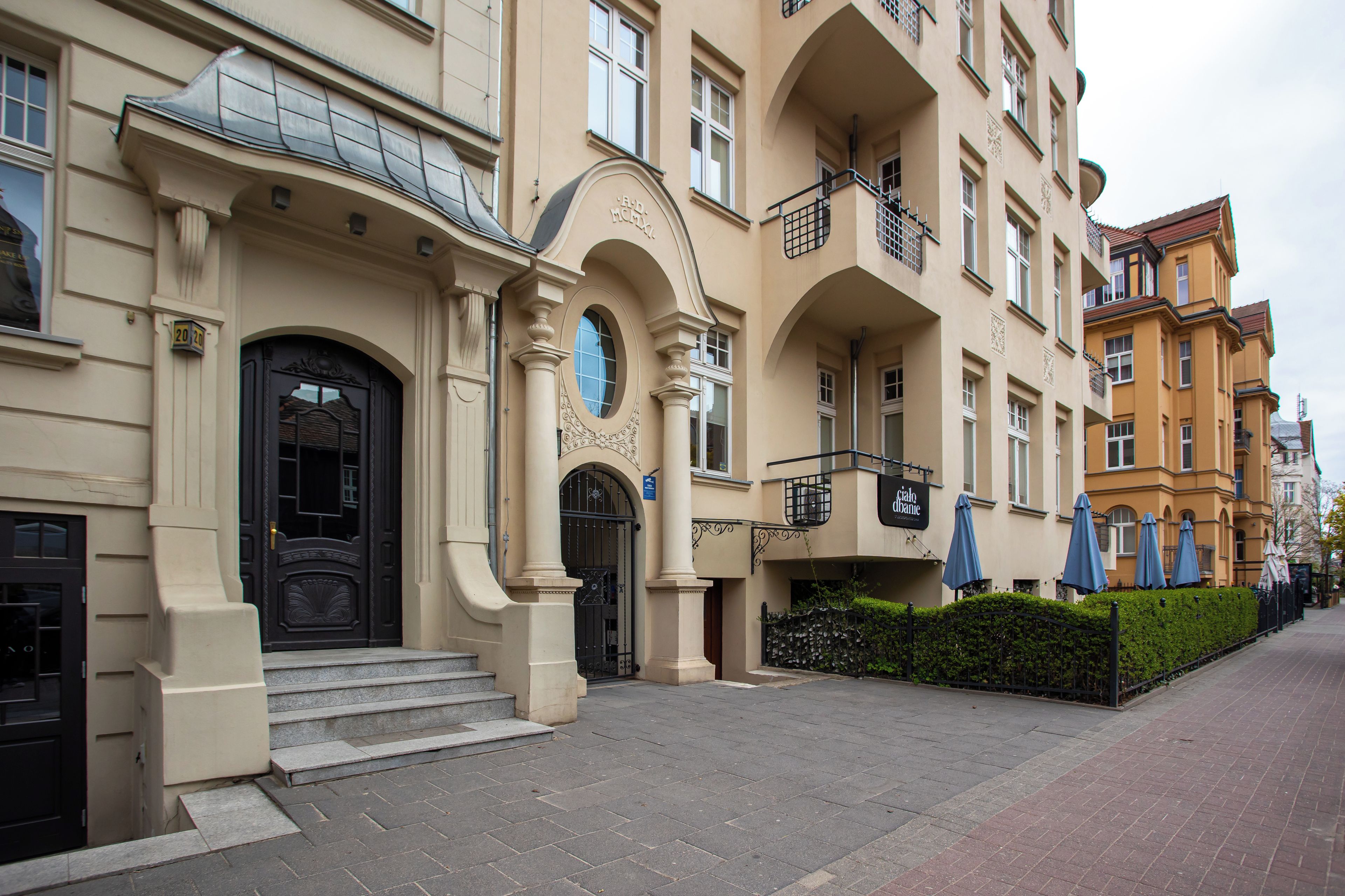 A multi-story building with a classic architectural style, featuring a prominent black entrance door, a balcony with a metal railing, and a sign indicating a restaurant.