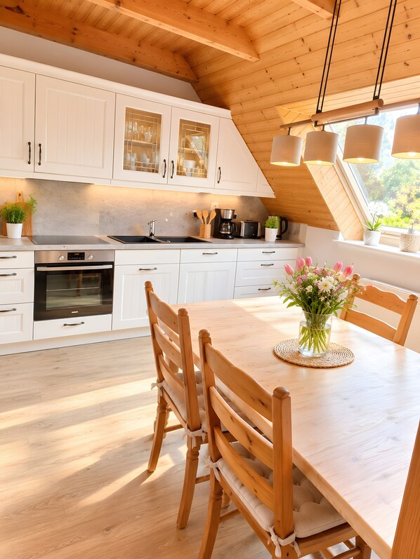 A modern kitchen with white cabinets, a wooden dining table, and a view of greenery outside.