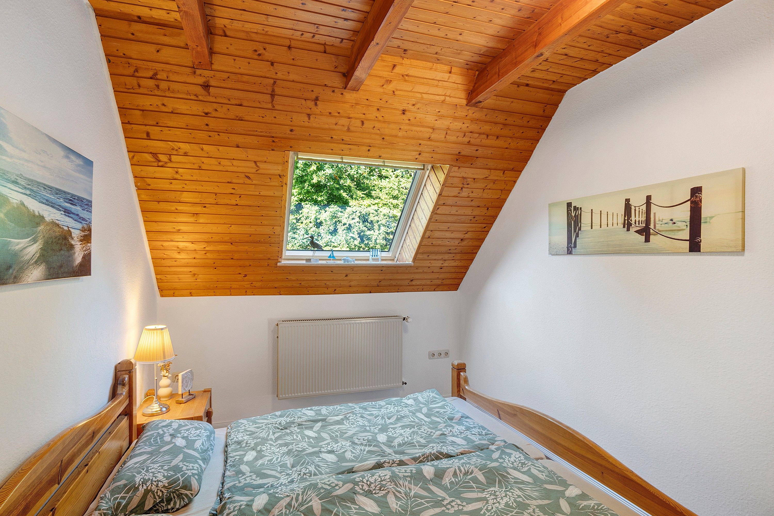Ein Schlafzimmer mit schrägen Wänden, einem Oberlicht, einem Bett mit floralem Bettzeug, einem Nachttisch mit Lampe und einer Heizung.