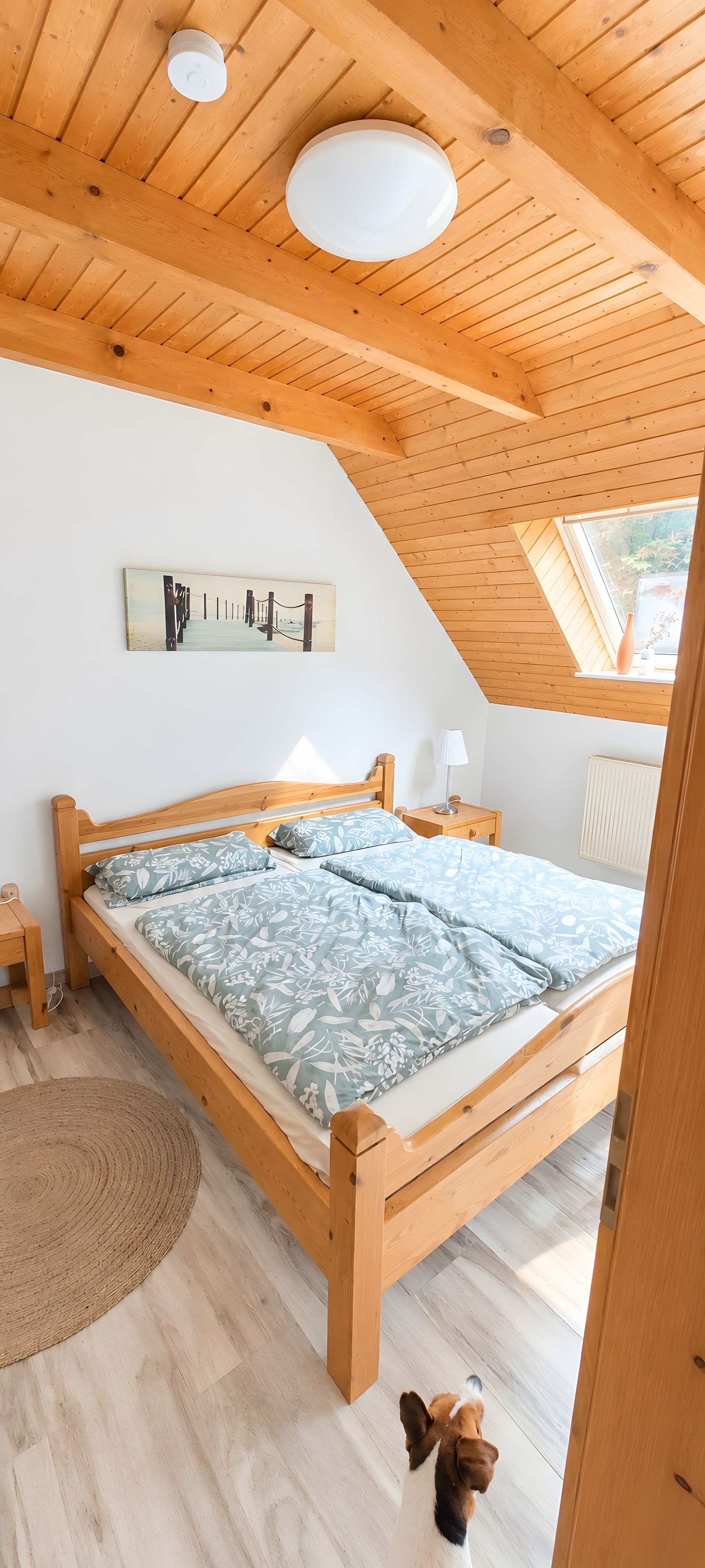 Ein Schlafzimmer mit Holzboden, einem Bett mit gemusterten Bettbezügen, einem Nachttisch, einer Lampe und einem Gemälde an der Wand.