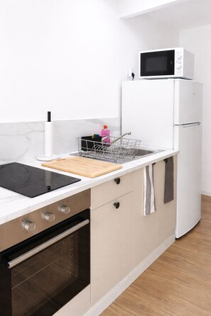 A kitchen with a black induction cooktop, a wooden cutting board, a stainless steel sink, a dish rack, a white refrigerator, and a microwave.