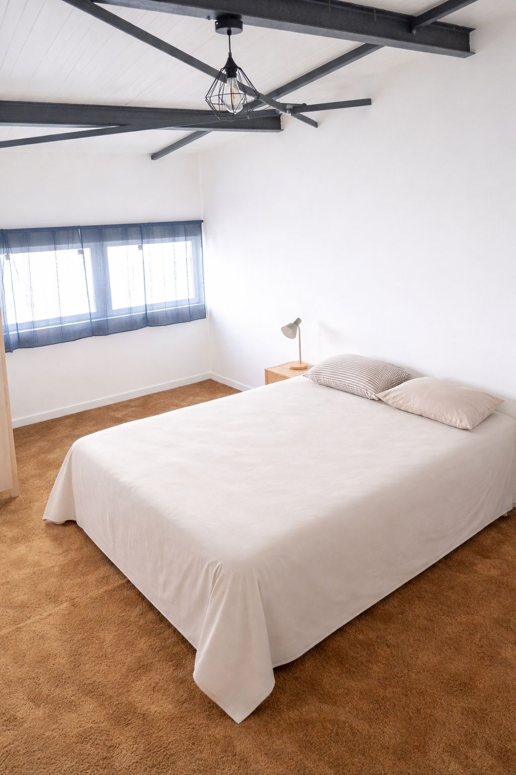 Un dormitori amb un llit gran, una tauleta de nit, una làmpada, una finestra amb cortines i un ventilador de sostre.