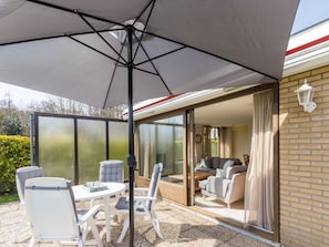 A patio area with a table and chairs, a large umbrella, and a sliding glass door leading to an interior space with a sofa and curtains. - Holiday Home in Renesse With Swimming Pool (Renesse)