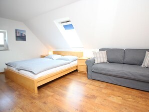 A bedroom with a bed, a grey sofa, a wooden nightstand, and a window with an air conditioning unit. - at the South Grove 3 (Sylt)