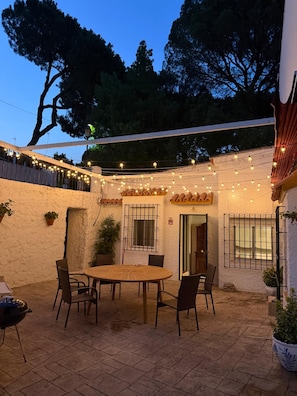 An outdoor patio area with a round table and chairs, string lights, and a white wall with a window.