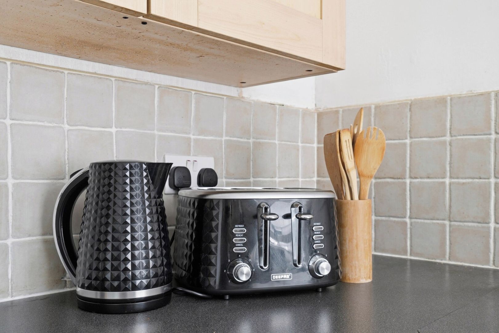 A kitchen counter with a black toaster, a black kettle, and a wooden utensil holder.