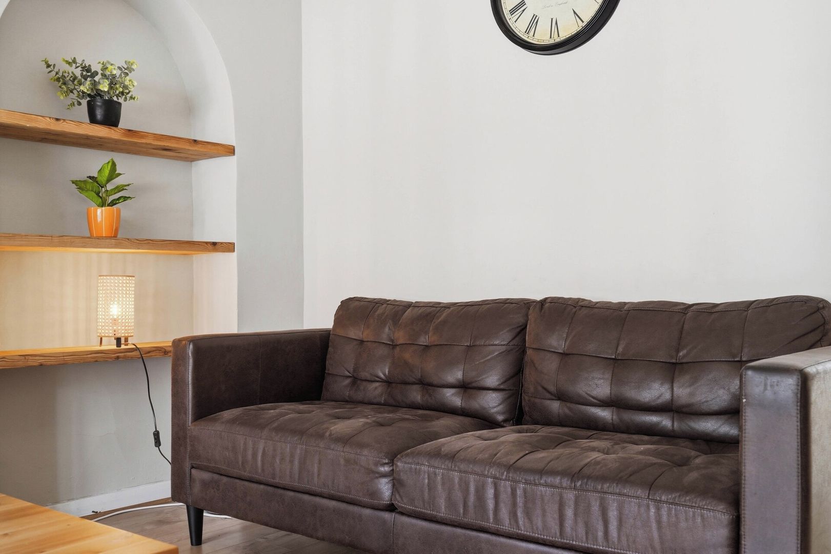Ein braunes Ledersofa in einem Raum mit Holzboden, einer Wanduhr und Regalen mit Pflanzen und einer Lampe.