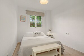 A bedroom with a bed, a nightstand, a lamp, a window with a view of greenery, and a framed map on the wall. - Apartment 'Baviera Golf Resort' with Shared Pool, Private Terrace and Wi-Fi (Caleta de Vélez)