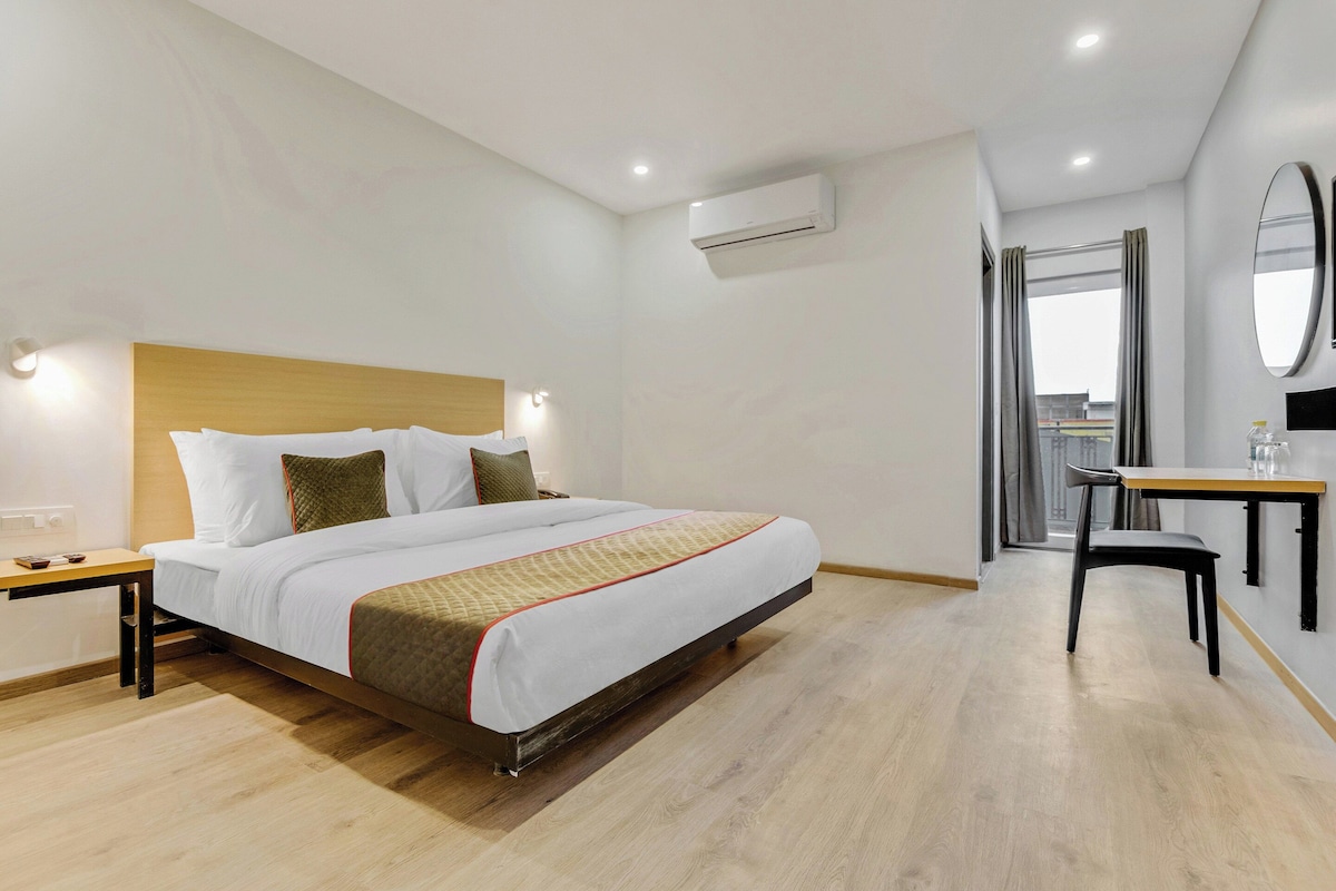 A hotel room with a bed, bedside tables, a desk with a chair, an air conditioning unit, and a window with curtains.