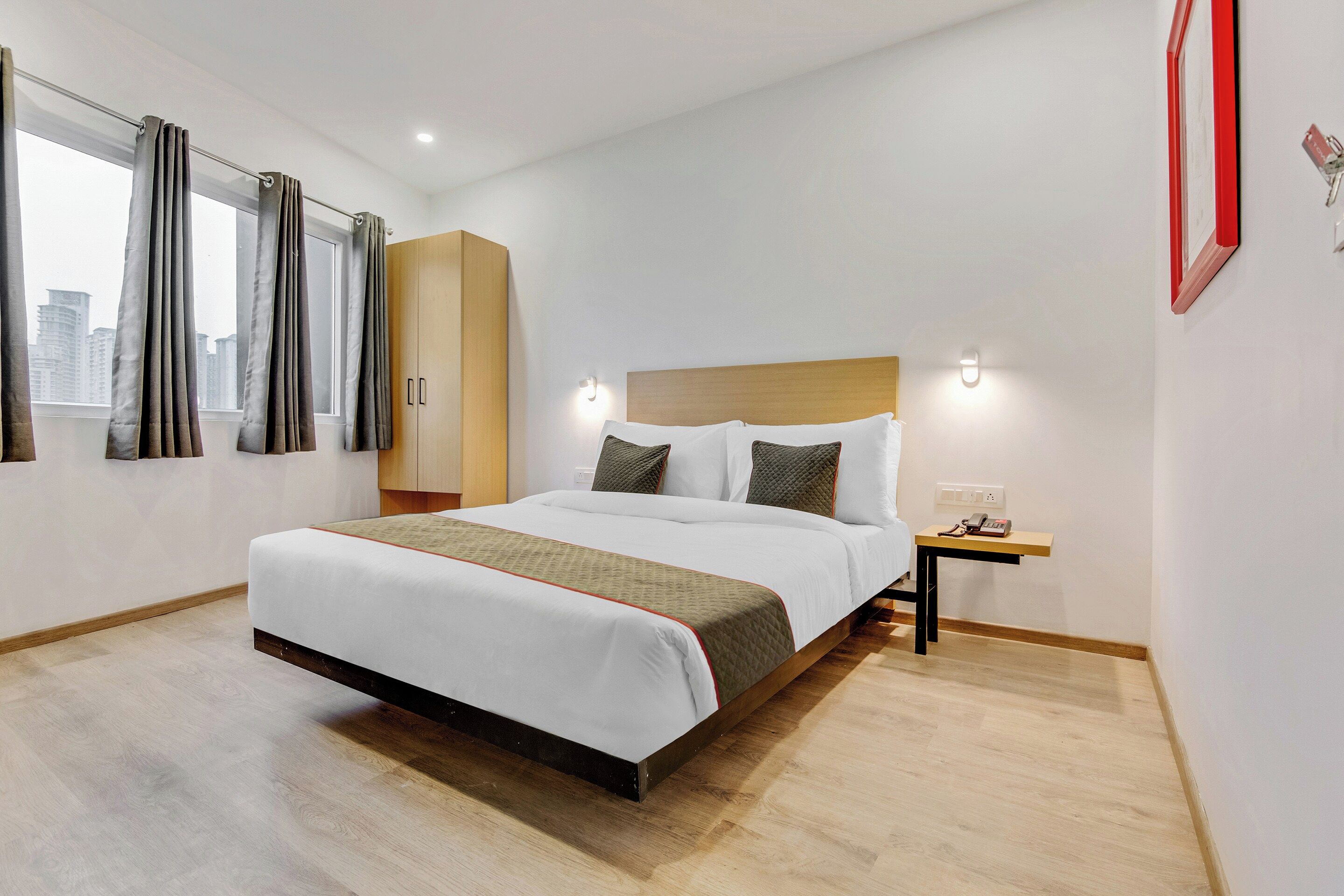 Une chambre d’hôtel avec un grand lit, une tête de lit en bois, une table de chevet avec un téléphone et une vue sur la ville par la fenêtre.