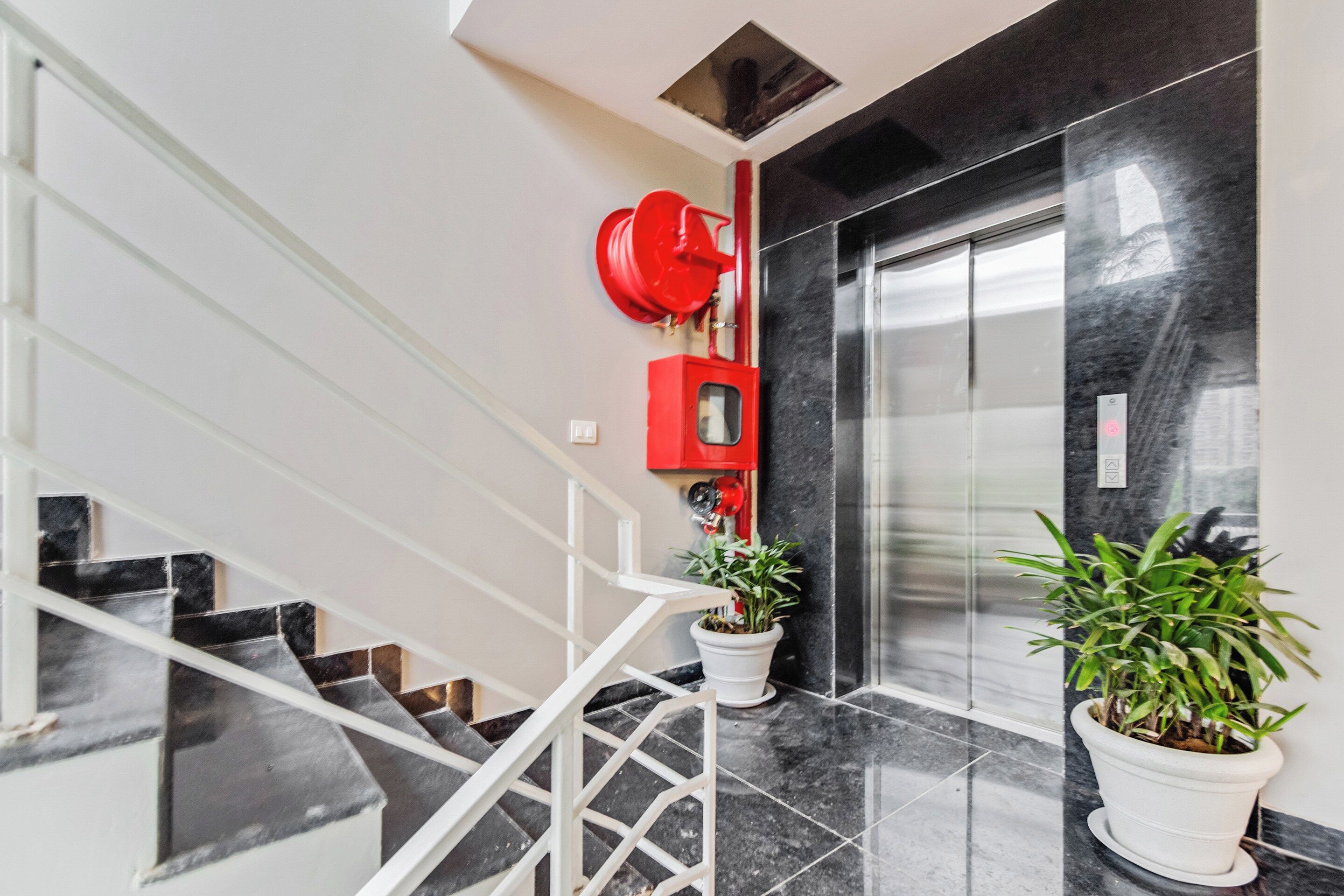 A modern interior with a glass elevator, a red fire alarm, and potted plants.