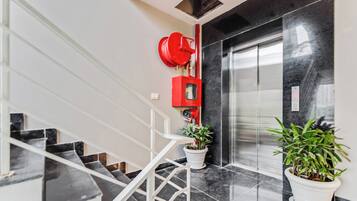 A modern interior with a glass elevator, a red fire alarm, and potted plants.