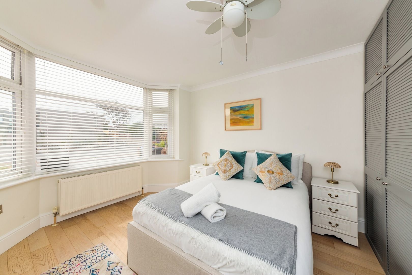Une chambre à coucher comprenant un lit, des tables de chevet, un radiateur, un ventilateur de plafond et une fenêtre avec des stores.