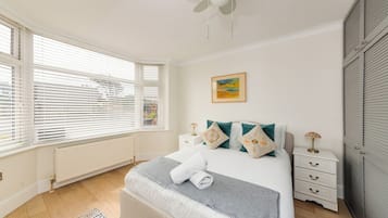 Une chambre à coucher comprenant un lit, des tables de chevet, un radiateur, un ventilateur de plafond et une fenêtre avec des stores.