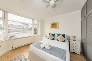A bedroom with a bed, bedside tables, a radiator, a ceiling fan, and a window with blinds. - Lancing 3BR Home | Parking for 3 Cars (West Sussex)