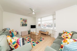A modern living room with a sofa set, a television, a ceiling fan, and a window with blinds. - Lancing 3BR Home | Parking for 3 Cars (West Sussex)