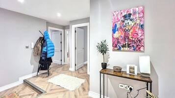 A hallway with a wooden floor, a coat rack with a blue jacket, a door, a wall-mounted plant, and a table with a power outlet.